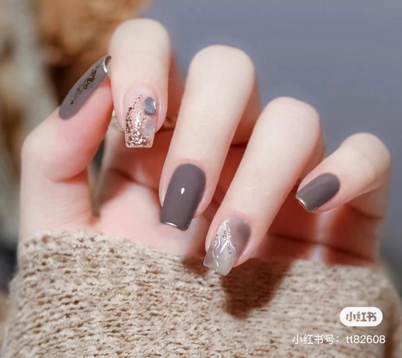 Bàn tay với móng tay sơn màu xám và trang trí họa tiết ánh kim, nền màu nâu nhạt.