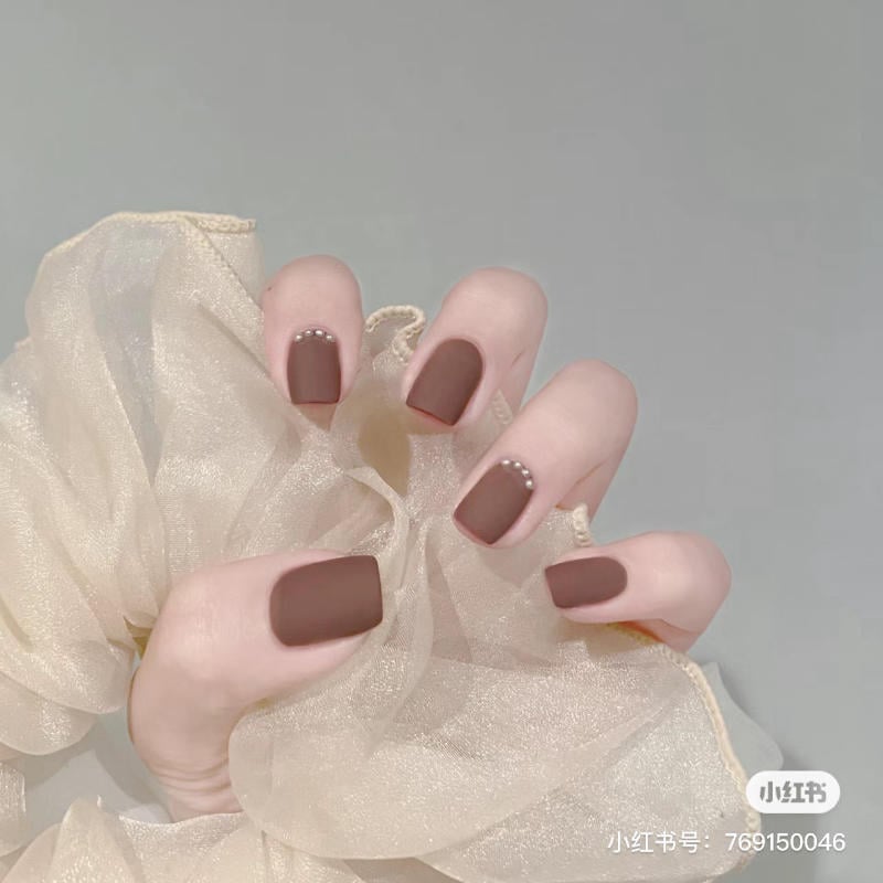 Bàn tay với móng tay sơn màu nâu đậm và đính đá, trên nền vải voan màu kem.