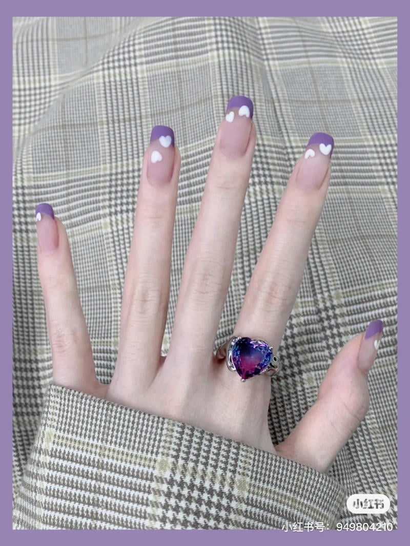 nail-mau-tim__10__231b83366f284e0eacd501601baf1032 Bàn tay với móng tay sơn đầu tím, họa tiết trái tim trắng, đeo nhẫn đá tím, trên nền vải caro.