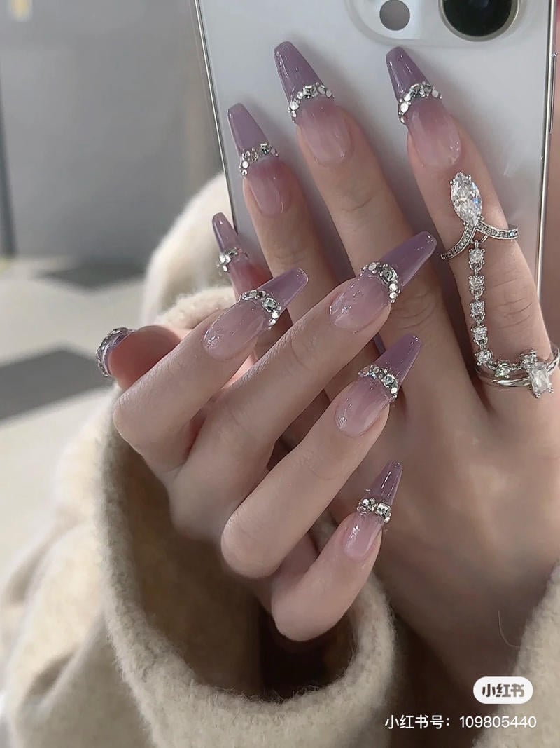 nail-mau-tim__12__263277d33fe64d169382c4225a29ad53 Tay với móng tay sơn màu tím và đính đá, cầm điện thoại, đeo nhẫn kim cương, áo khoác lông.