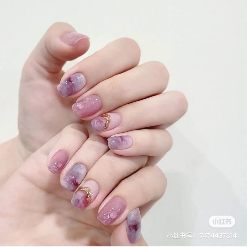 nail-mau-tim__15__2f5ab5ccc0f24b78a823754977e98508 Bàn tay với móng tay được trang trí màu hồng tím, họa tiết và chi tiết ánh vàng, nền trắng.