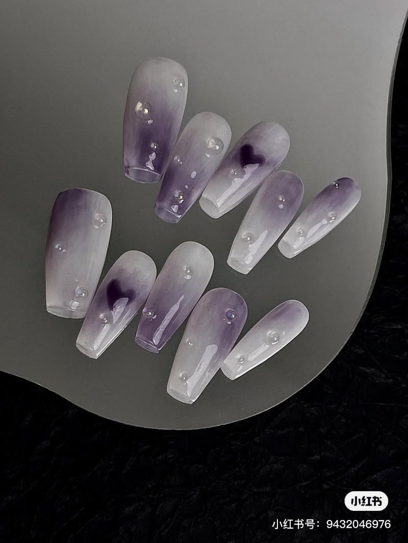 nail-mau-tim__16__86b71f13ec434fcfbaf205121cafe7c1 Bộ móng tay giả màu tím nhạt với họa tiết giọt nước, đặt trên nền đen.