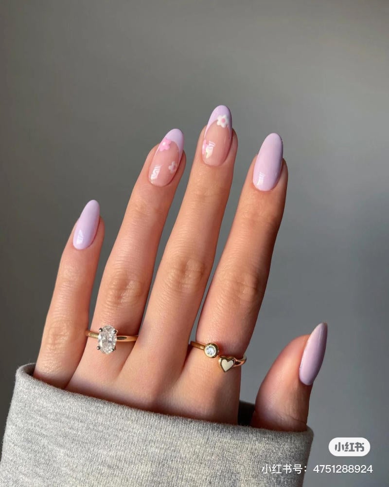 nail-mau-tim__17__e98b47e152ad4469be70778485f96a96 Tay với móng tay sơn màu tím nhạt và họa tiết hoa, đeo nhẫn vàng, nền xám.