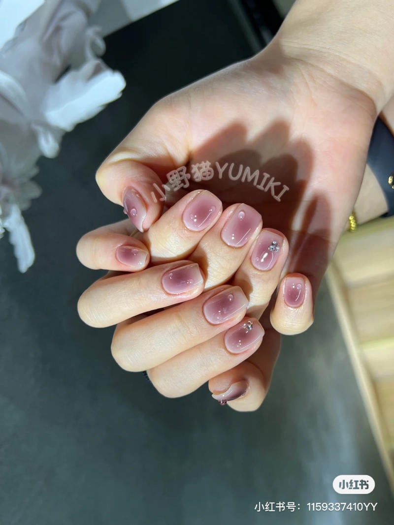 nail-mau-tim__20__1855c3c7f8ce49d09b232ad5f2ccdb71 Bàn tay với móng tay được sơn màu hồng nhạt và trang trí đá nhỏ, nền tối.