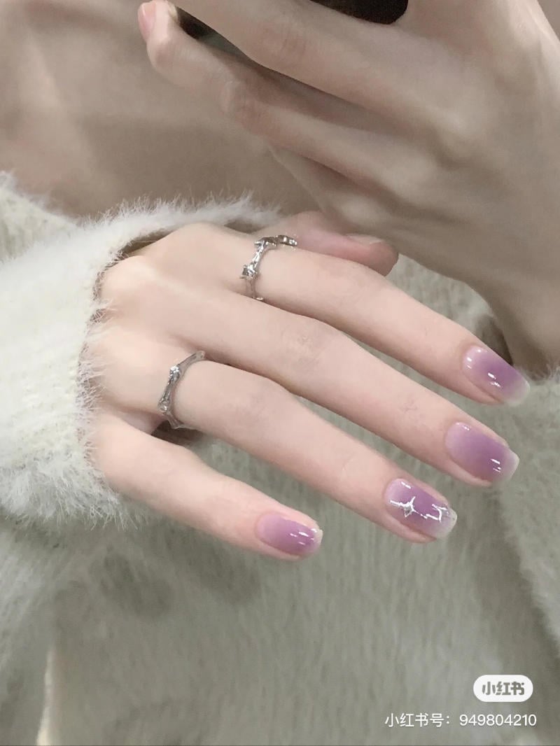 nail-mau-tim__24__33252e8adc704bffbfd5489bc7694516 Bàn tay với móng tay ombre tím và họa tiết trang trí, đeo nhẫn bạc, cầm điện thoại.