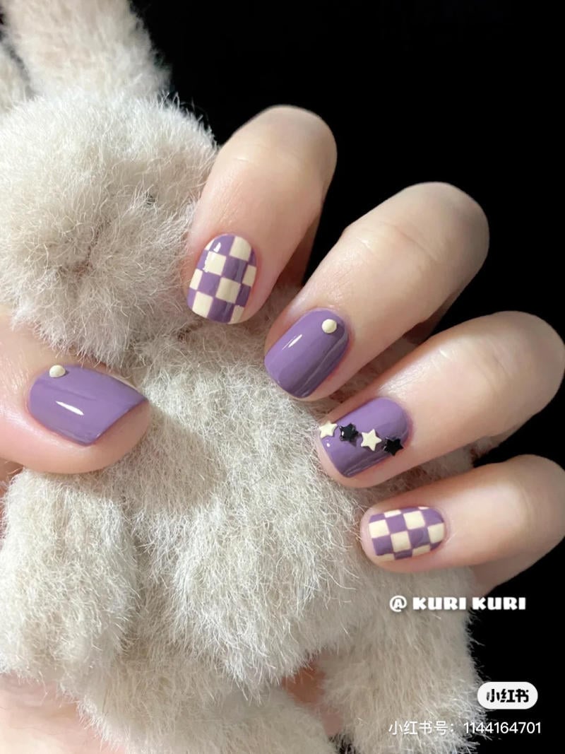 nail-mau-tim__25__97292f88282f4a5097069cf9c8e7022c Bàn tay với móng tay màu tím trang trí hoa văn caro và sao, cầm một vật liệu mềm màu trắng.