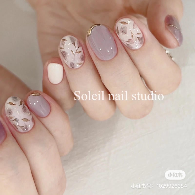 nail-mau-tim__26__978c0974c5cb41a587954fb8713cb093 Bàn tay với móng tay được sơn nghệ thuật, tông màu tím và trắng, họa tiết hoa lá và ánh kim.