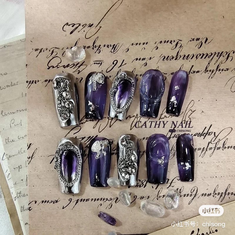 nail-mau-tim__28__c12216bfb7dc4115b13def0aa490cd77 Bộ móng giả màu tím với họa tiết kim loại phức tạp, nằm trên nền giấy viết tay cổ điển.
