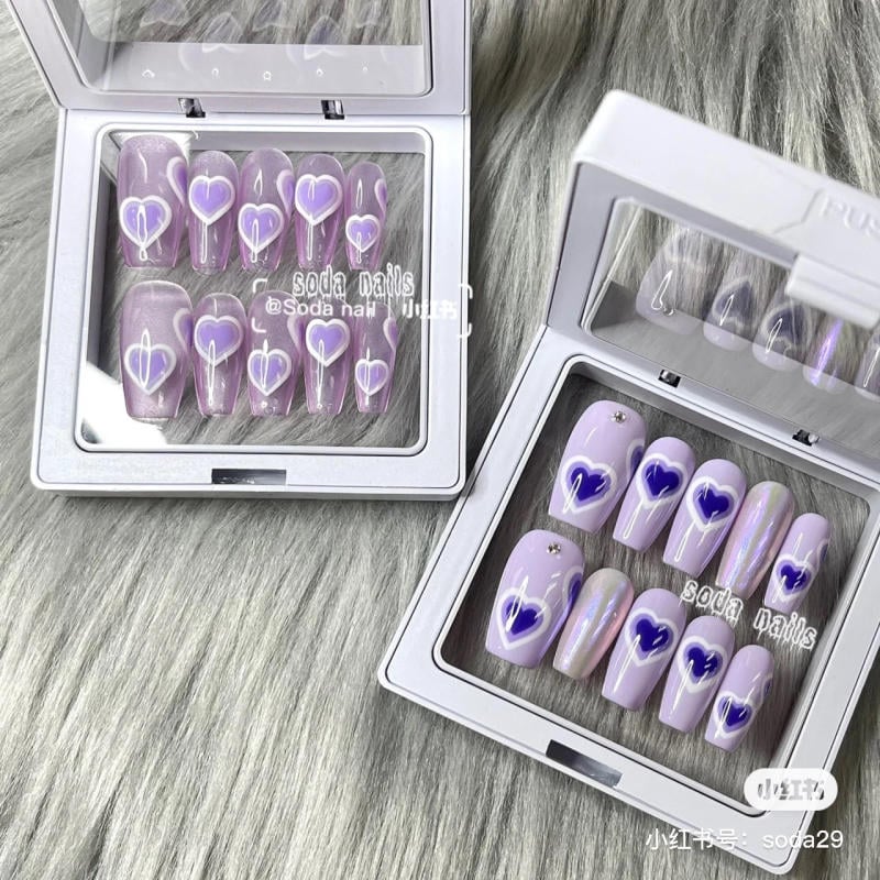 nail-mau-tim__2__666d1ae5215a4db080980737c2a15bb6 Hộp móng tay giả màu tím với họa tiết trái tim trắng và xanh, đặt trên nền lông xám.