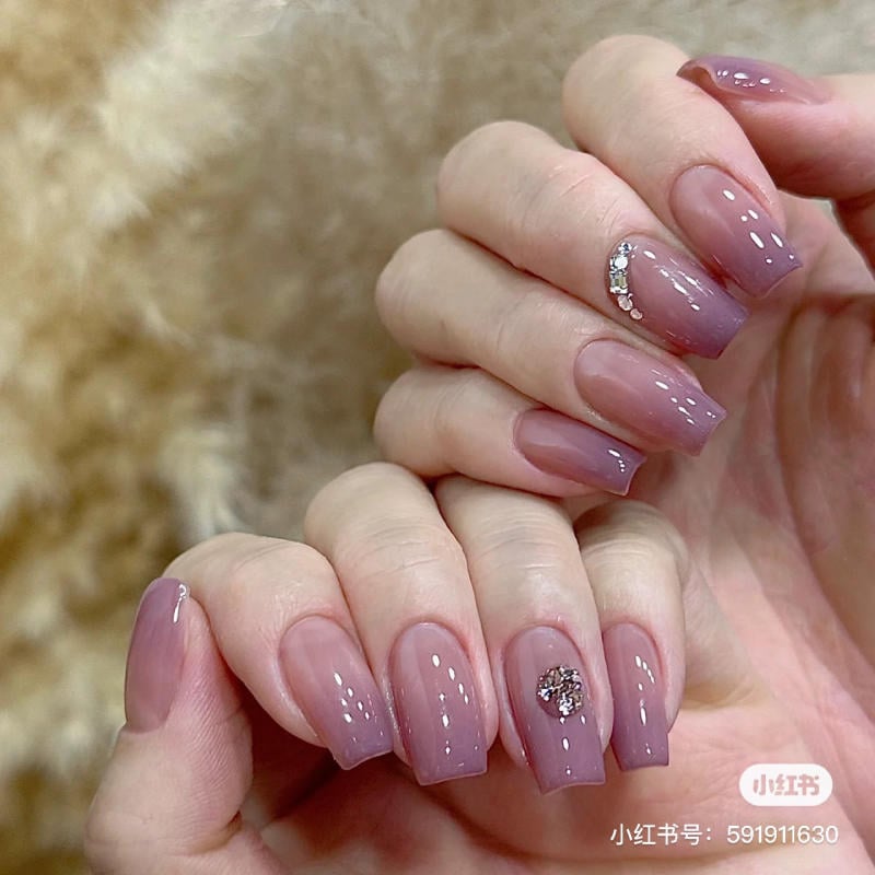 nail-mau-tim__35__ec03feeb94204c8ba1a91d44374c2e87 Bàn tay với móng tay màu tím nhạt, trang trí đá lấp lánh trên nền mờ, nền lông mềm mại phía sau.