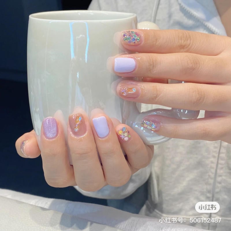 nail-mau-tim__39__f29a0209d76642ba97546875addbe39e Bàn tay với móng tay sơn màu pastel và trang trí cầm tách sứ trắng ở nền mờ.