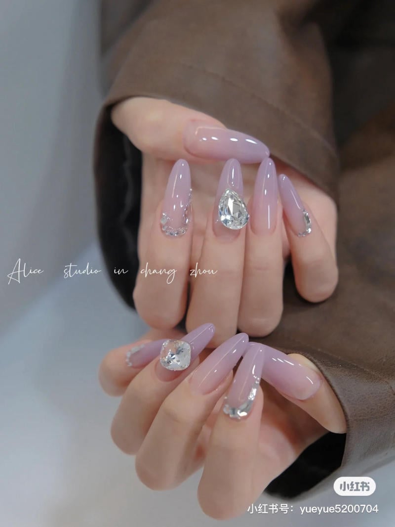 nail-mau-tim__43__790908b7354b481e93a458b854bd0213 Bàn tay với móng dài, sơn màu tím nhạt, đính đá sáng lấp lánh, tạo vẻ đẹp sang trọng và tinh tế.