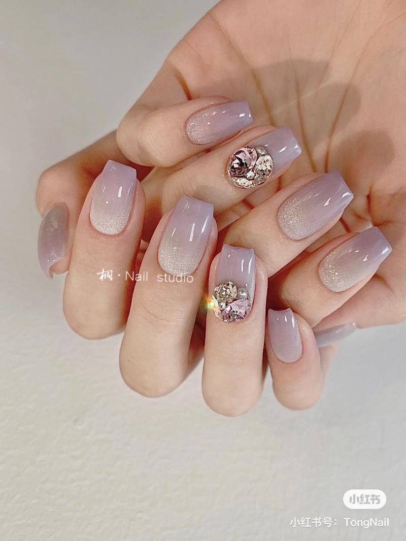 nail-mau-tim__44__da4d8125019042408ff38d6c22cbe204 Bàn tay với móng tay được sơn màu tím nhạt, có trang trí đính đá lấp lánh trên một ngón.