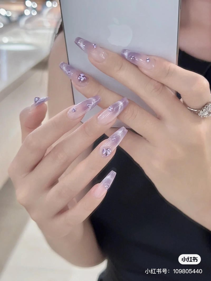 nail-mau-tim__46__ca2a641d168140e592fa7d7a2fa76f32 Người cầm điện thoại, móng tay dài màu tím nhạt, đính đá, trên nền môi trường nội thất sáng.
