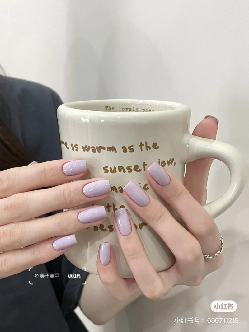 nail-mau-tim__49__a351f0ce3acc4e0f86b030d3131e0c71 Đôi tay với móng tay màu tím nhạt cầm tách sứ màu trắng có chữ trang trí.