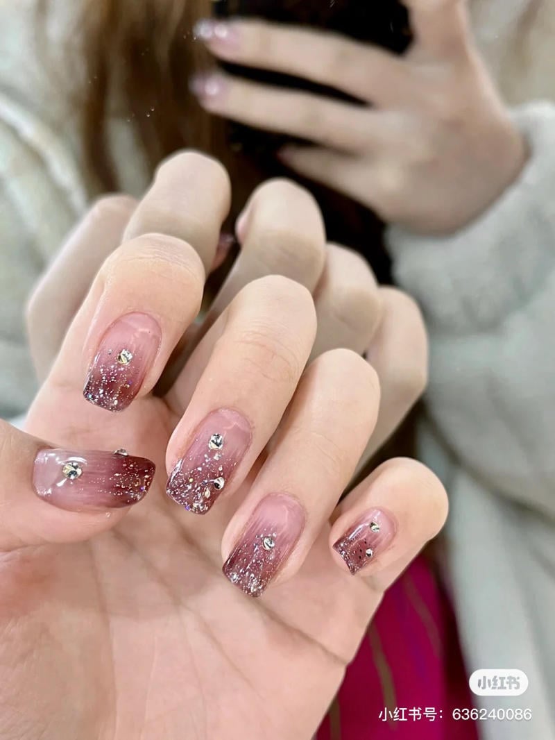 nail-mau-tim__55__152784933fd04fec8607acfffc079217 Bàn tay với móng tay nghệ thuật màu hồng nhạt, đính đá lấp lánh và họa tiết lấp lánh.