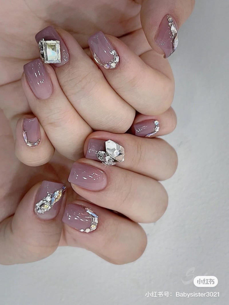 nail-mau-tim__58__d95d91301b274c7d9dc1f06498ba6f52 Bàn tay với móng tay màu tím nhạt, trang trí đá lấp lánh và họa tiết kim loại.
