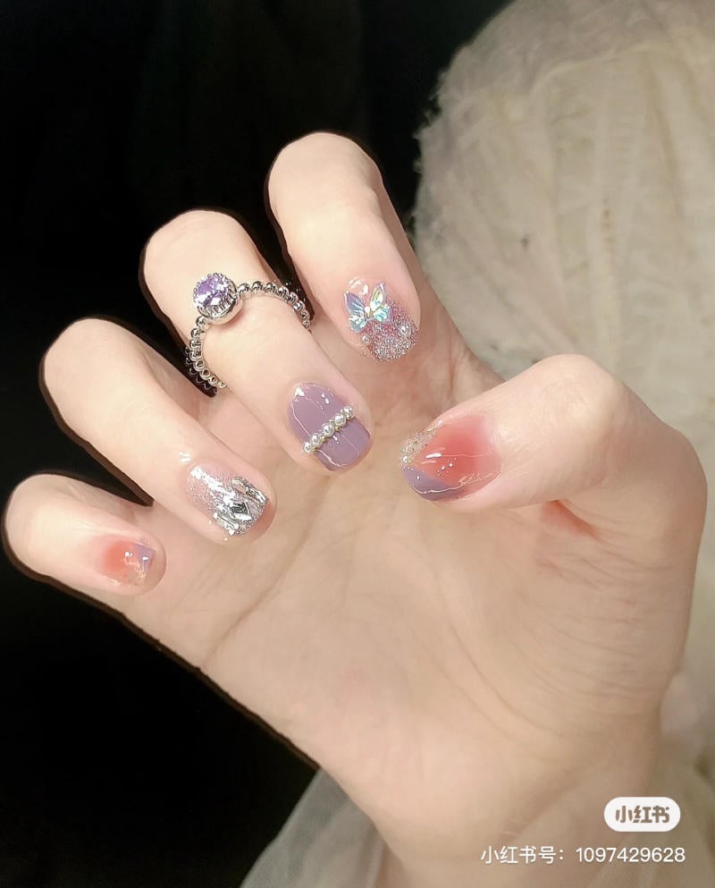 nail-mau-tim__5__03cc50b1945b4ccea398f27f8f83cfc6 Bàn tay với móng tay được trang trí cầu kỳ, có nhẫn và họa tiết lấp lánh trên nền tối.