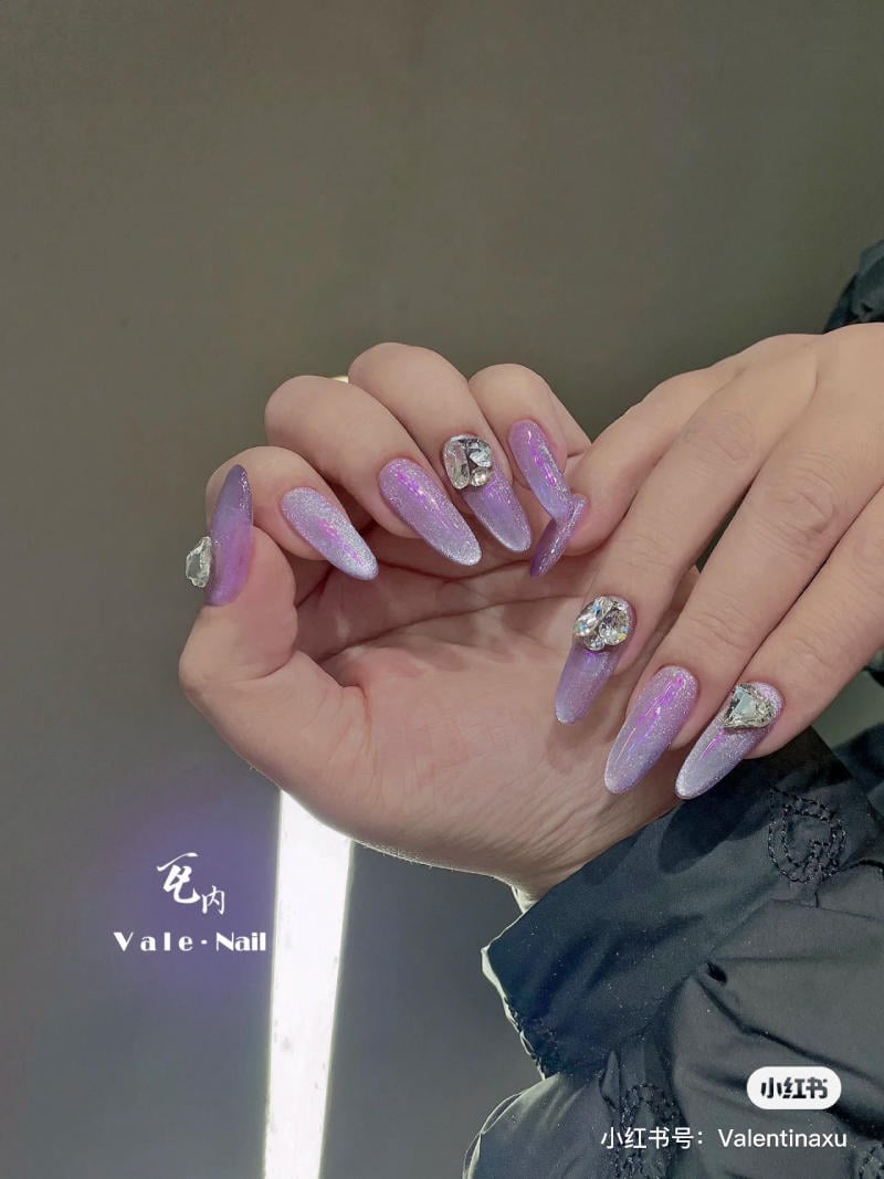 nail-mau-tim__70__6cd72221c5364974bf48a7fe76b0a28c Bàn tay với bộ móng dài màu tím óng ánh, gắn đá lấp lánh, trên nền xám.