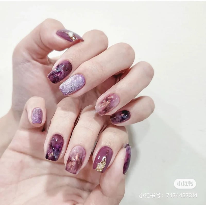 nail-mau-tim__71__83113583c95d479181af6e26b77c1796 Bàn tay với móng tay được sơn màu tím, trang trí hoa văn và đính phụ kiện nhỏ.