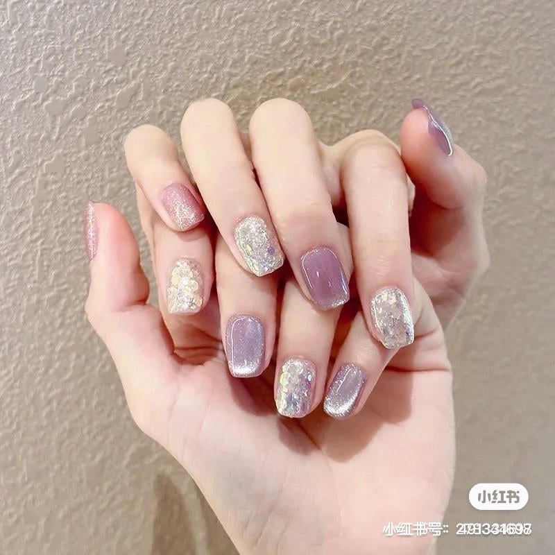 nail-mau-tim__74__b4b76888e29142d3b3d583303e3d320e Bàn tay với móng tay được sơn màu tím nhạt và trang trí bằng kim tuyến lấp lánh, chụp trên nền tường sáng.