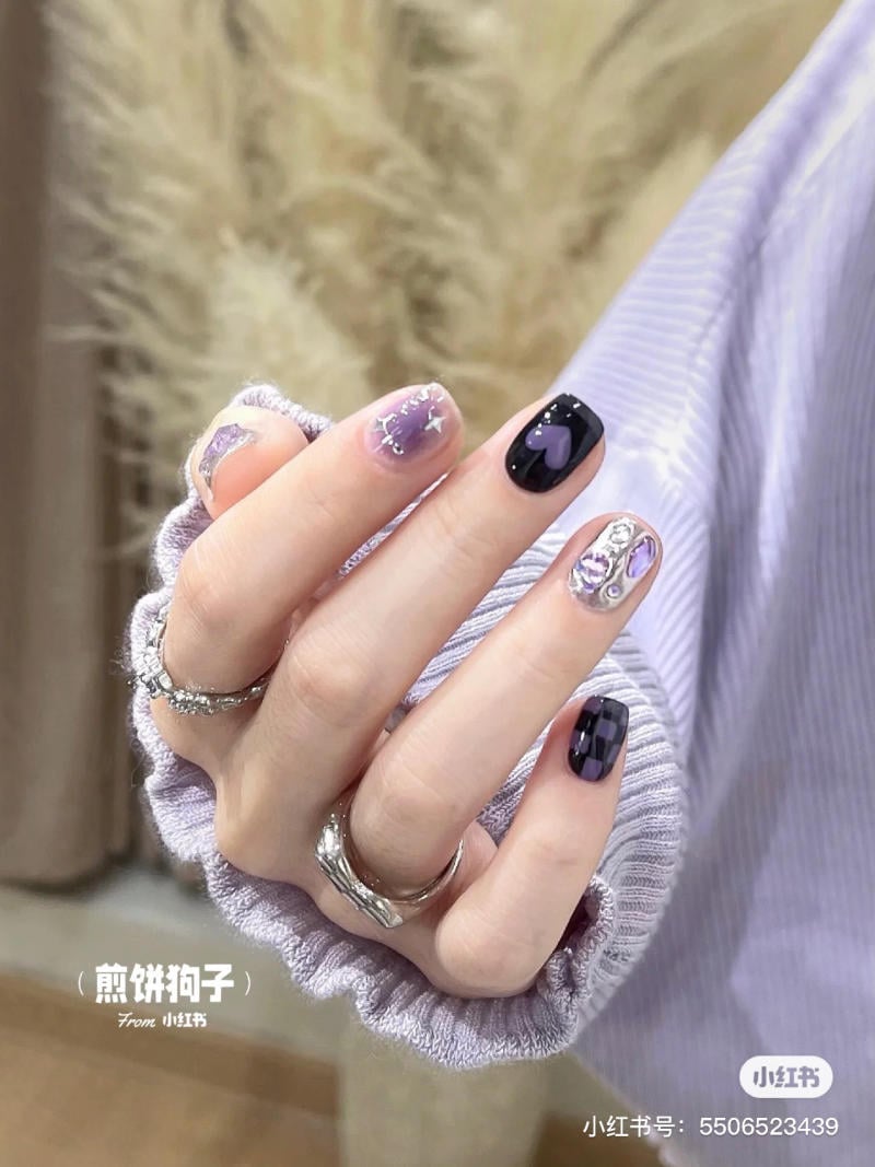 nail-mau-tim__7__1373241d909d461088986529969f15da Bàn tay với móng tay đính đá và thiết kế hoa văn màu tím, đen. Tay đeo nhẫn bạc và áo tay dài màu tím nhạt.