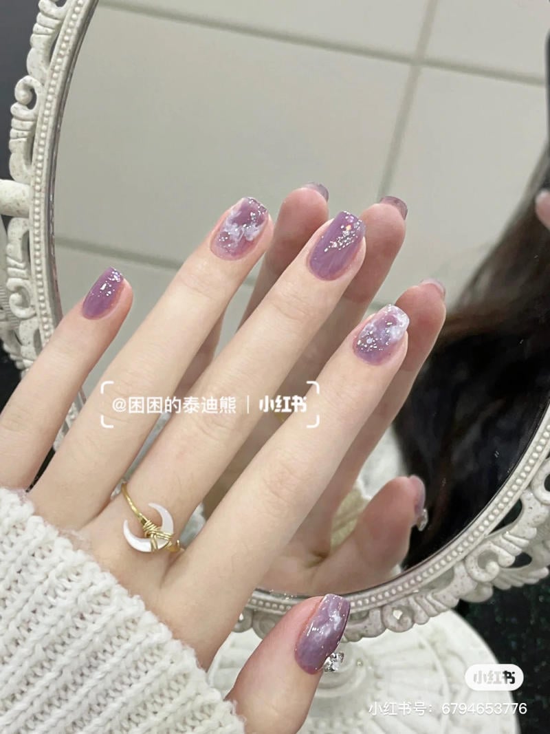 nail-mau-tim__82__309f88f0c54247e985e662c658be9e0a Tay với móng sơn màu tím nhạt, trang trí họa tiết, đeo nhẫn hình mặt trăng, phản chiếu trong gương viền hoa văn.