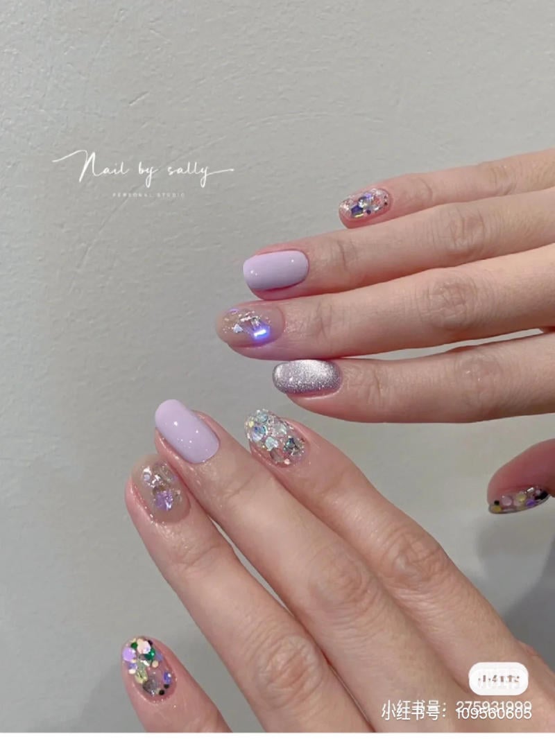 nail-mau-tim__88__48e117d3381a4066997e55a643e2f4e0 Bàn tay với móng tay được sơn màu tím nhạt và trang trí kim tuyến lấp lánh trên nền màu nhạt.