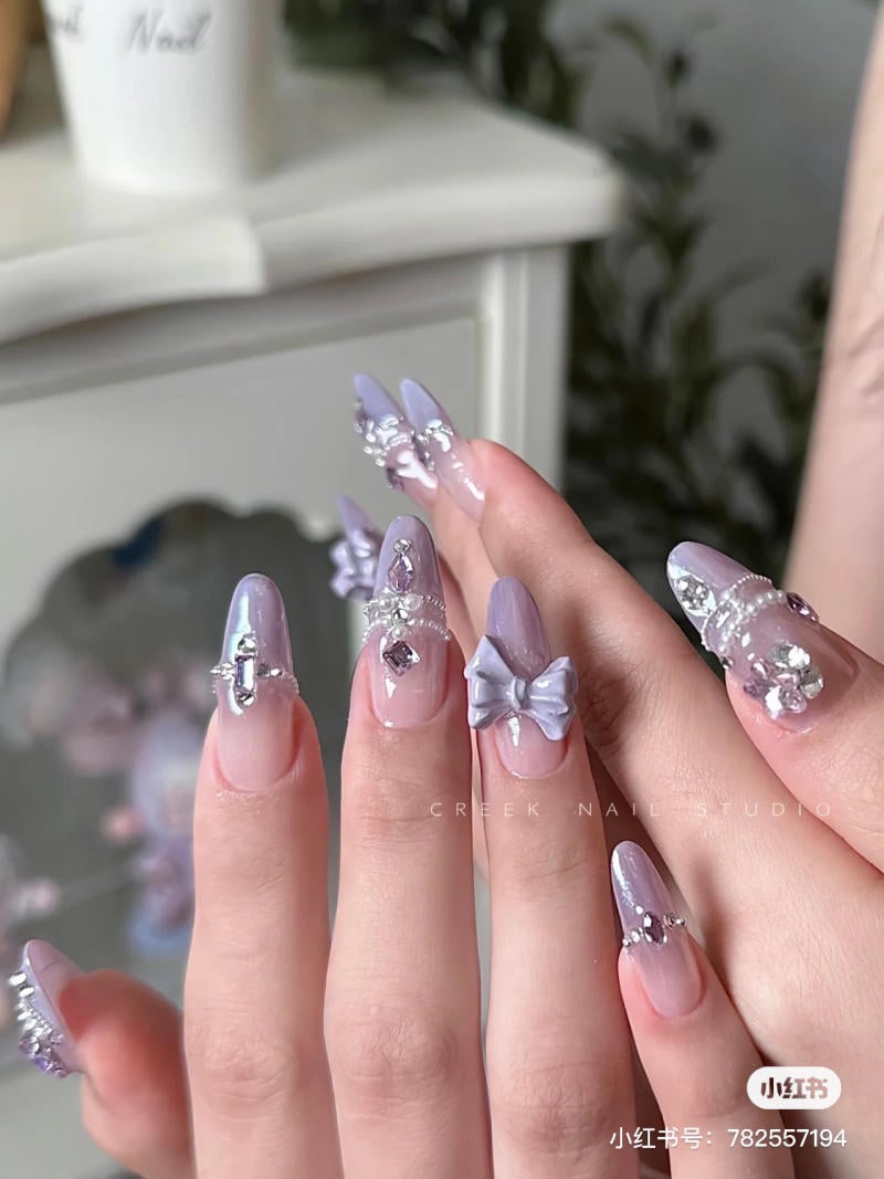 nail-mau-tim__8__13a103aef35c48f386139a1939c1badc Bàn tay với móng tay dài được trang trí bằng màu tím nhạt và phụ kiện hình nơ cùng đá lấp lánh.