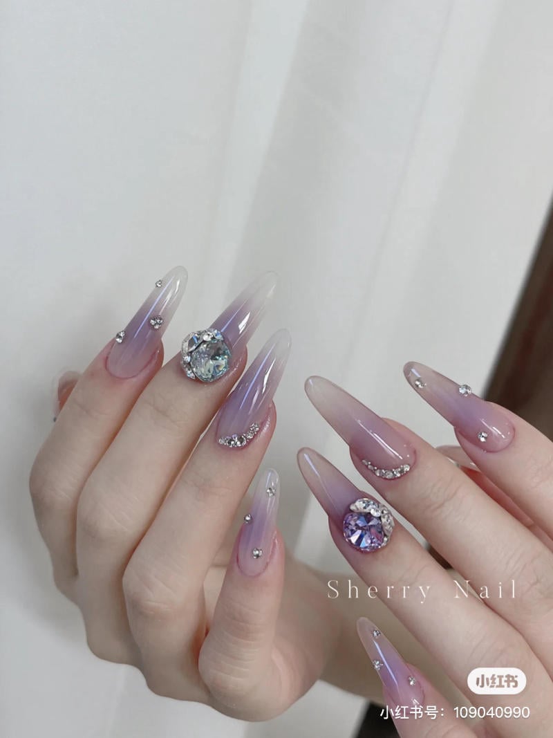 nail-mau-tim__9__49eb813ff875494ebb131cca5eb97f68 Bàn tay với móng tay dài sơn màu ombre tím hồng, trang trí đá lấp lánh và họa tiết tinh xảo.