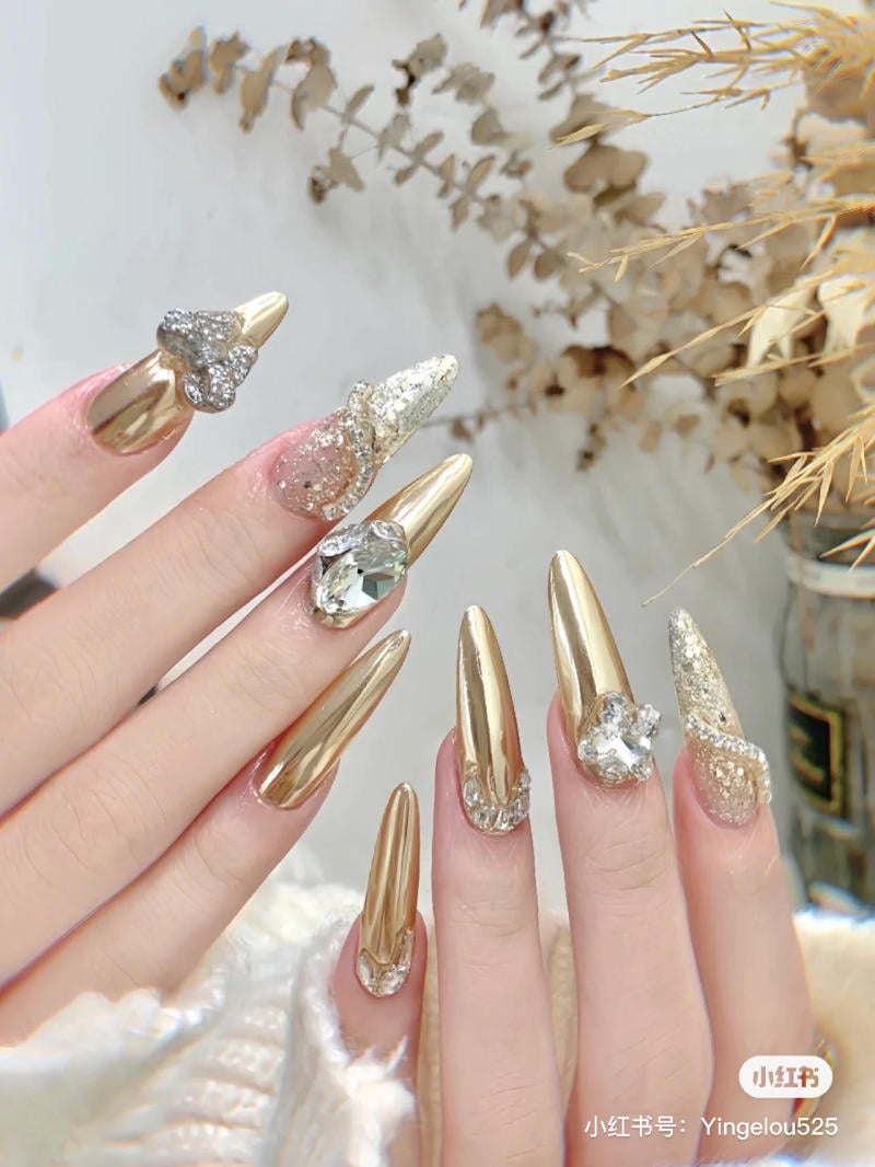 Bàn tay với móng tay dài, được sơn màu vàng bóng và đính đá, nền có cây khô trang trí.