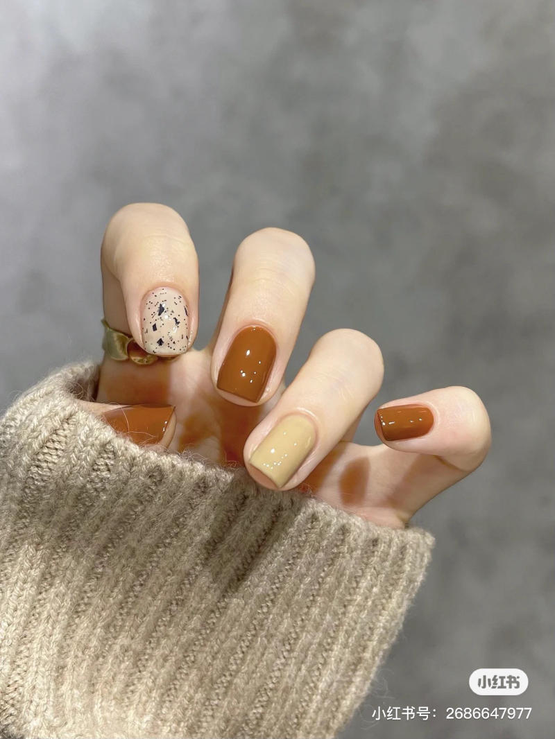 Bàn tay đeo nhẫn với móng tay sơn màu nâu, be, và họa tiết, mặc áo len màu nâu nhạt.