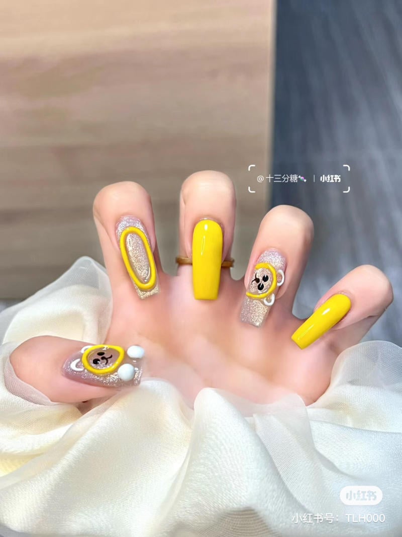 Bàn tay với móng tay được trang trí màu vàng và hình gấu, kết hợp với các chi tiết ánh kim lấp lánh.