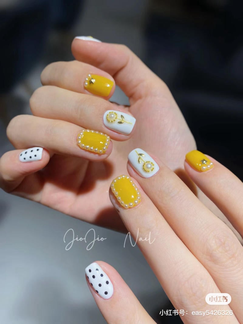 Bàn tay với móng tay được sơn màu vàng và trắng, có hoa văn hoa và chấm bi đen.