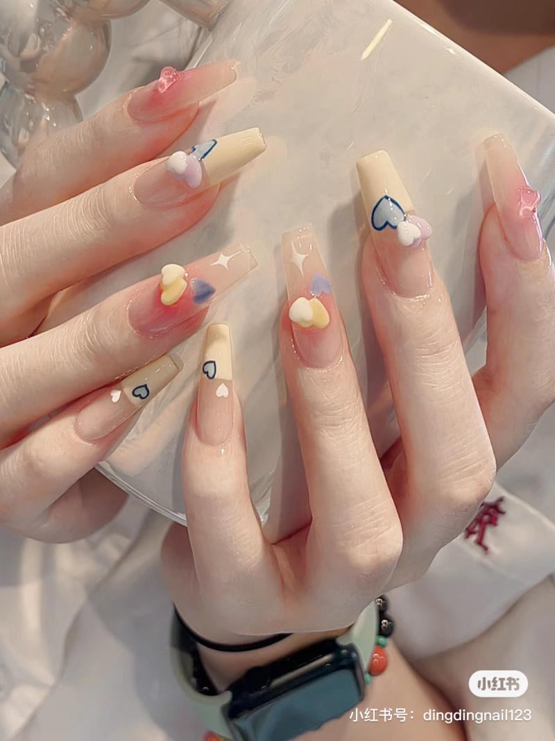 Bàn tay với móng tay dài được trang trí bằng họa tiết hình trái tim nhiều màu sắc trên nền móng màu nude.