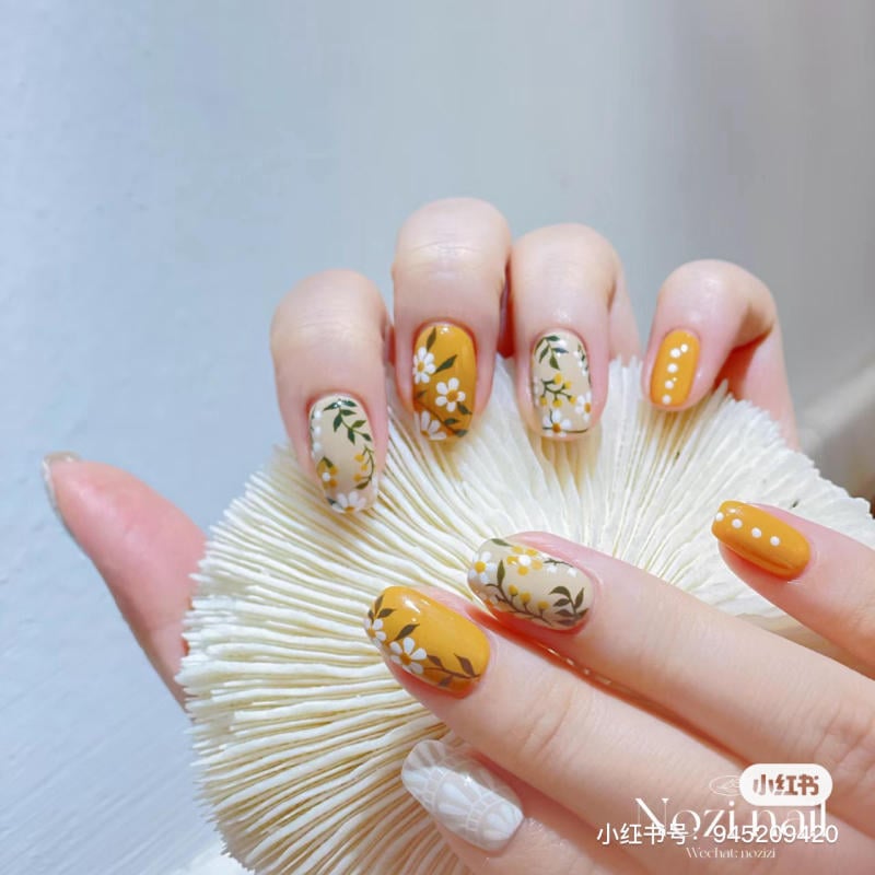 Bàn tay với móng nghệ thuật màu vàng và trắng, họa tiết hoa và chấm tròn, cầm vật thể màu trắng.