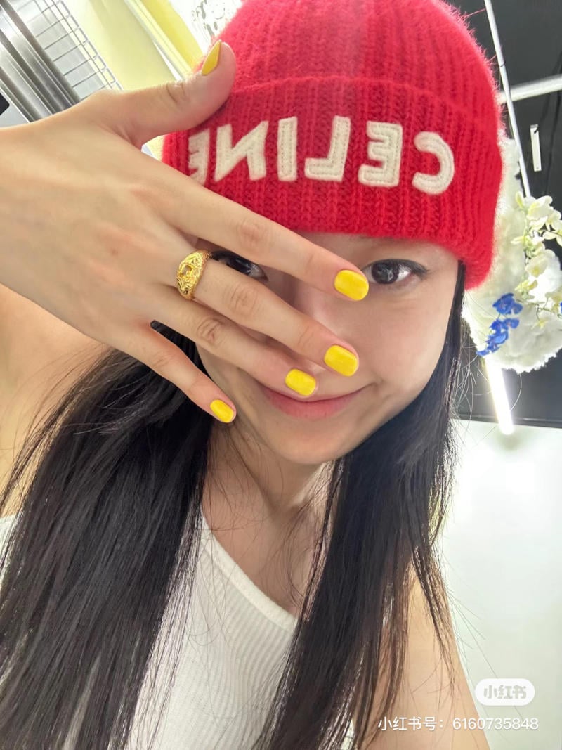 Người đội mũ len đỏ, giơ tay có móng sơn vàng và đeo nhẫn vàng, mỉm cười trước ống kính.