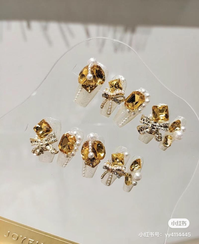 Bộ móng giả trang trí đá quý màu vàng và ngọc trai, thiết kế cầu kỳ và sang trọng trên nền trắng.