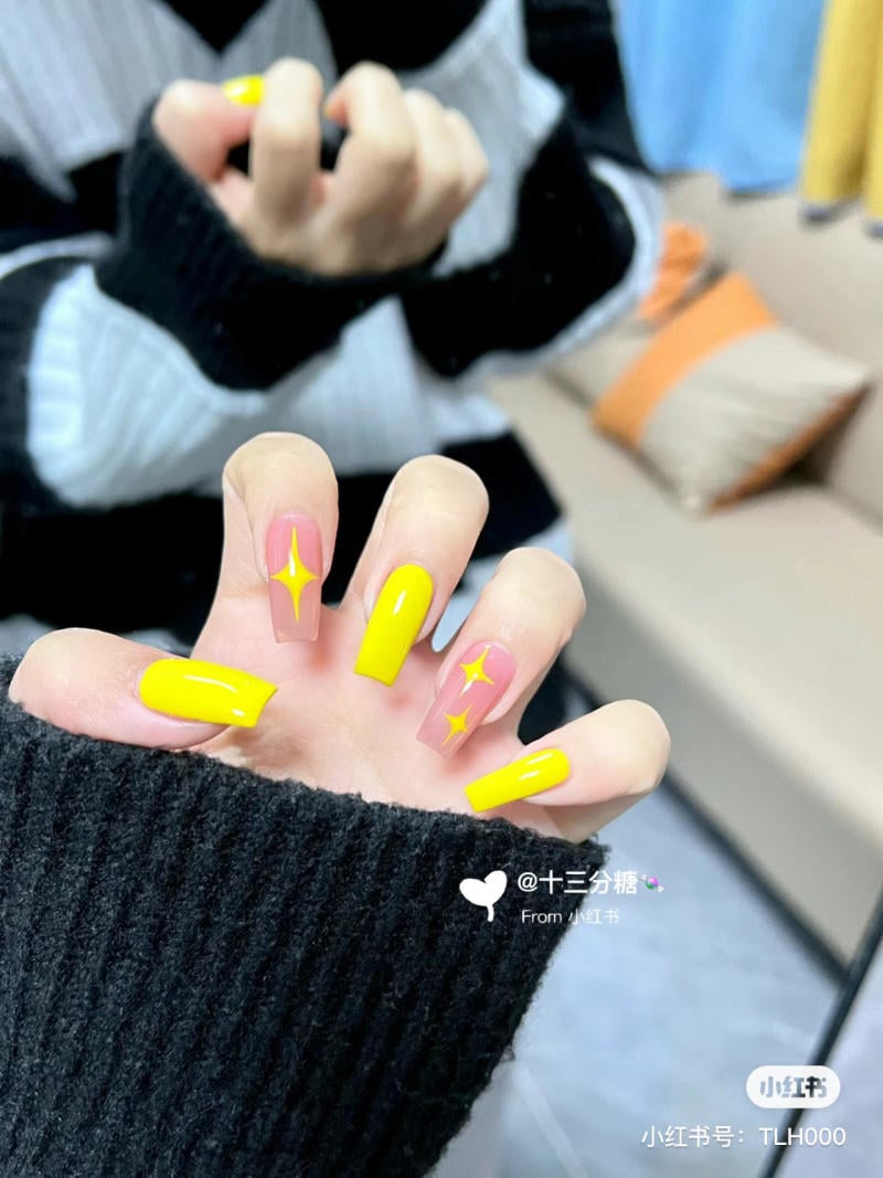 Bàn tay với móng tay sơn màu vàng và họa tiết ngôi sao, mặc áo len đen trắng, nền là ghế sofa.