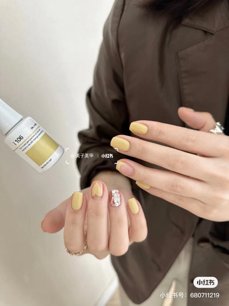 Bàn tay với móng tay sơn màu vàng, một ngón có họa tiết đính đá, cạnh chai sơn móng tay cùng màu.