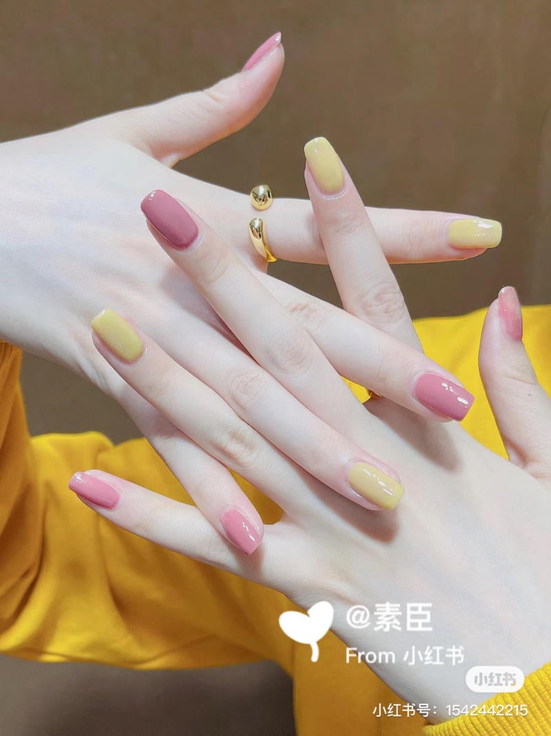 Bàn tay với móng tay sơn màu vàng và hồng, cùng một chiếc nhẫn vàng, trên nền áo vàng.