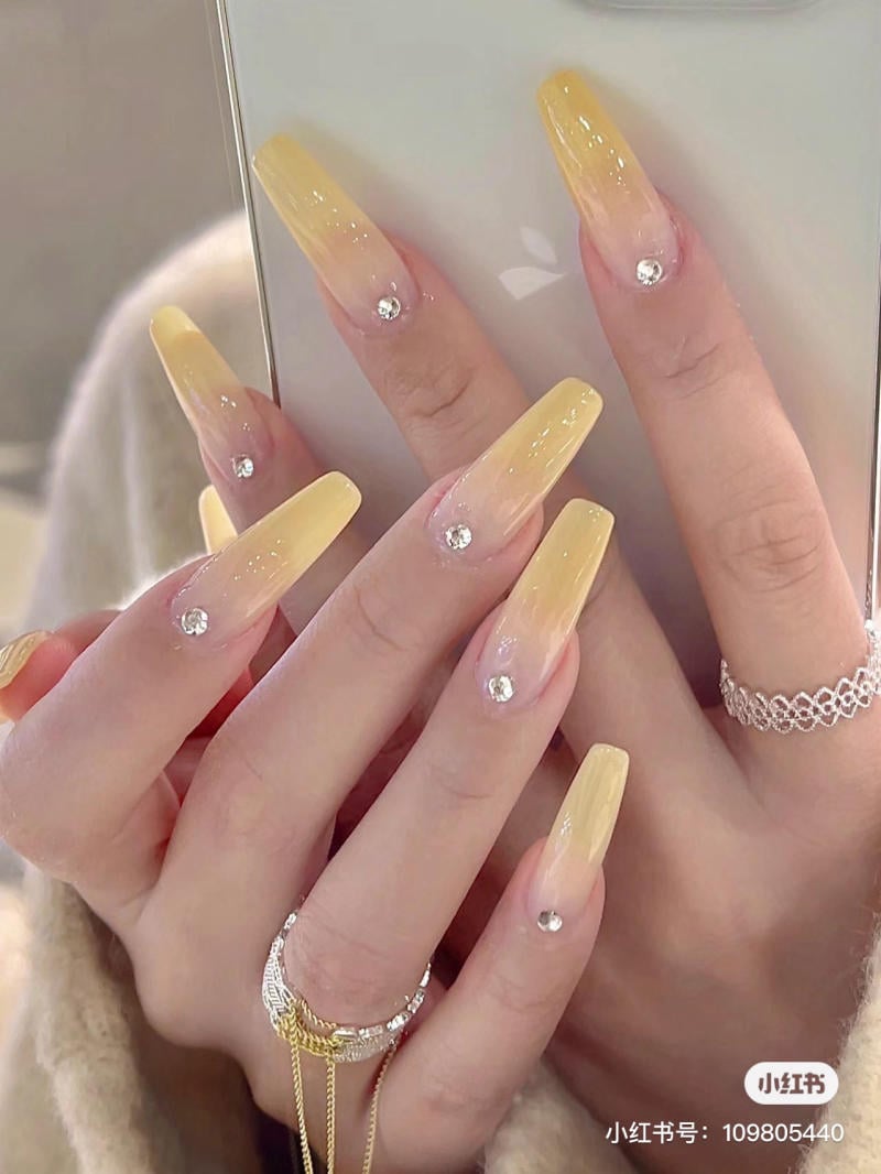 Bàn tay với móng tay dài màu vàng, đính đá, cầm điện thoại. Ngón tay có đeo nhẫn vàng và bạc.