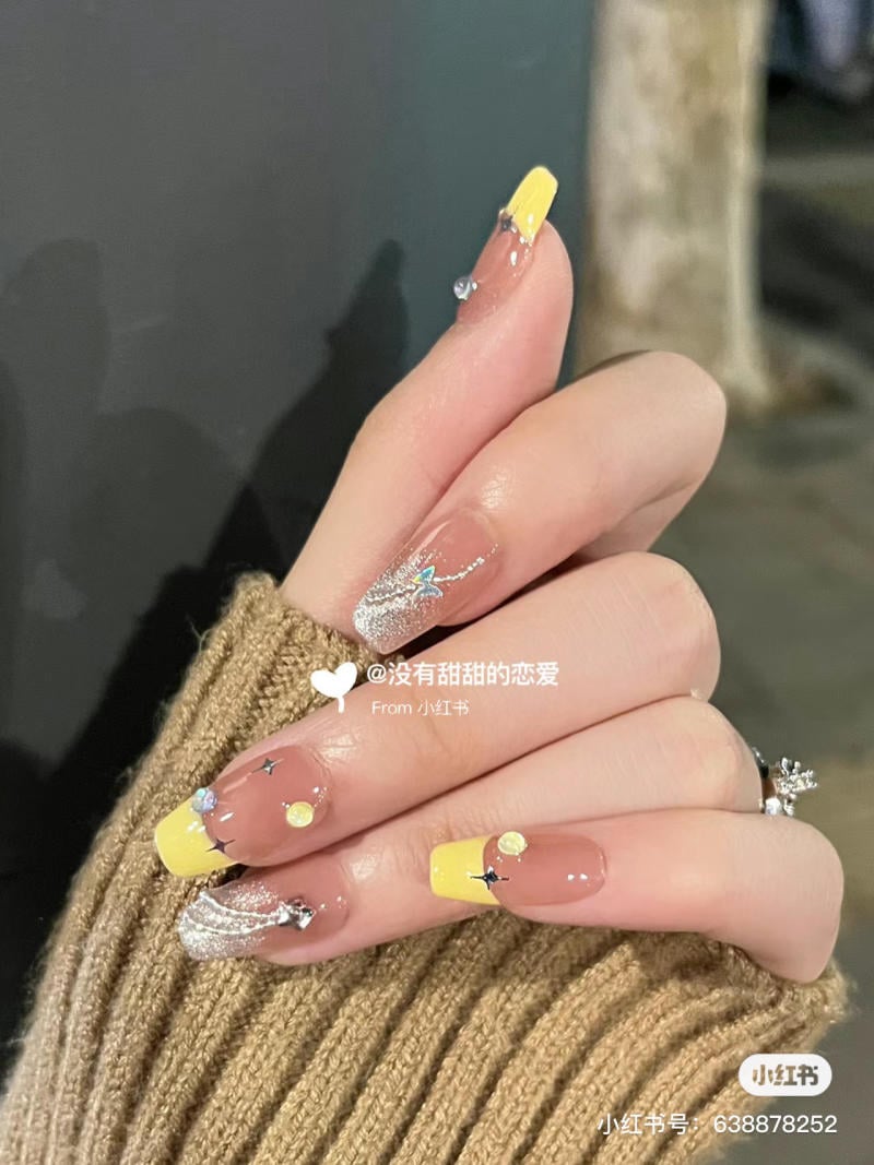 Bàn tay với móng tay được sơn màu vàng và trang trí họa tiết lấp lánh, trên nền áo len màu nâu.