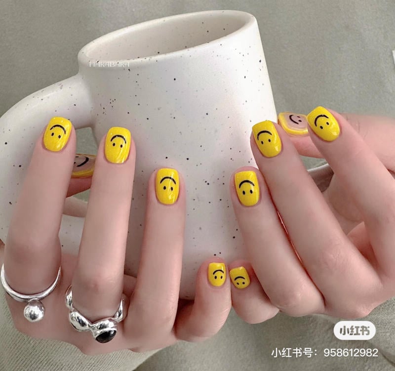 Bàn tay với móng sơn màu vàng và họa tiết mặt cười, cầm một chiếc cốc trắng có vết đốm nhỏ.