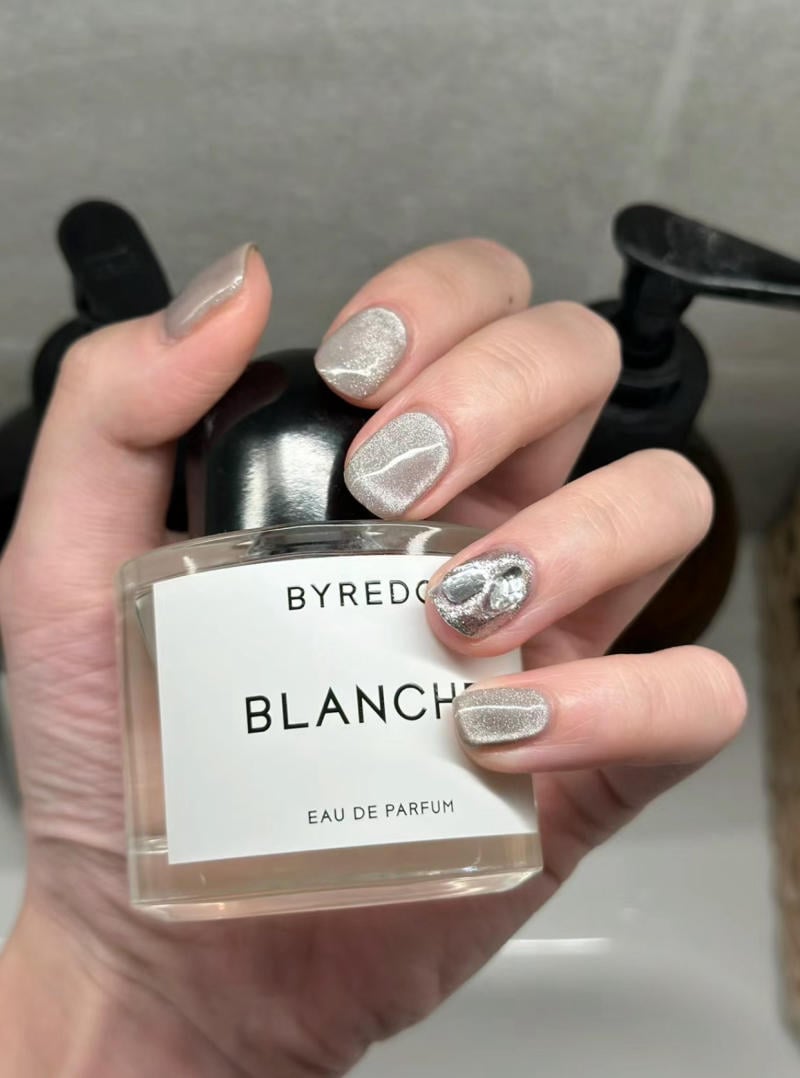 Tay với móng tay sơn bạc cầm chai nước hoa Blanche, nền phông xám.