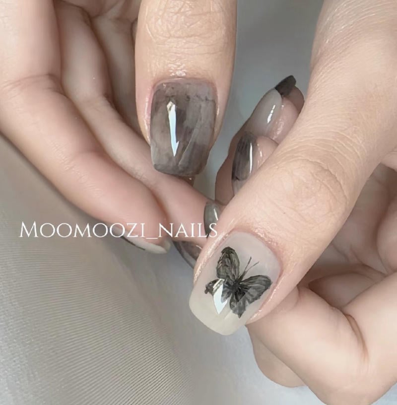 Bàn tay với móng tay sơn màu xám và một móng có hình vẽ bướm đen trên nền màu nhạt.