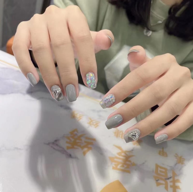 Bàn tay với móng tay sơn màu xám và trang trí lấp lánh, đặt trên bàn có hoa văn chữ Trung Quốc.