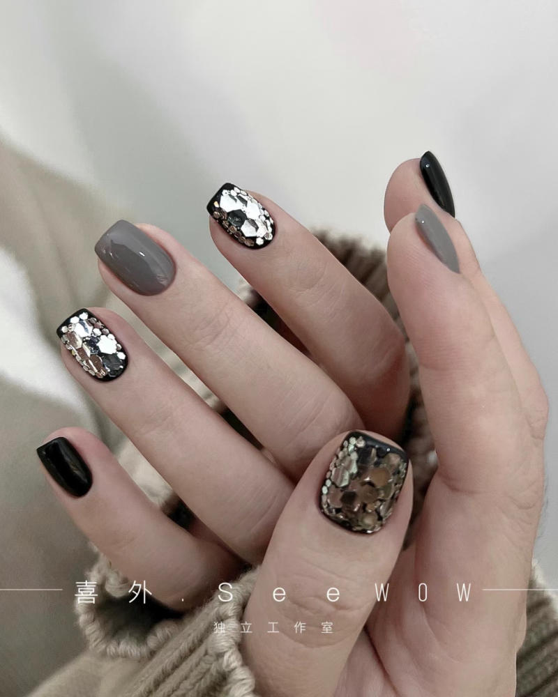 Bàn tay với móng tay được sơn màu đen, xám và đính kim loại lấp lánh, tạo kiểu nghệ thuật nổi bật.