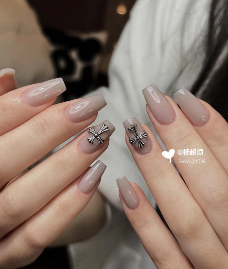 Bàn tay với móng tay dài màu nude, hai móng trang trí nơ kim loại sáng bóng, chụp cận cảnh.