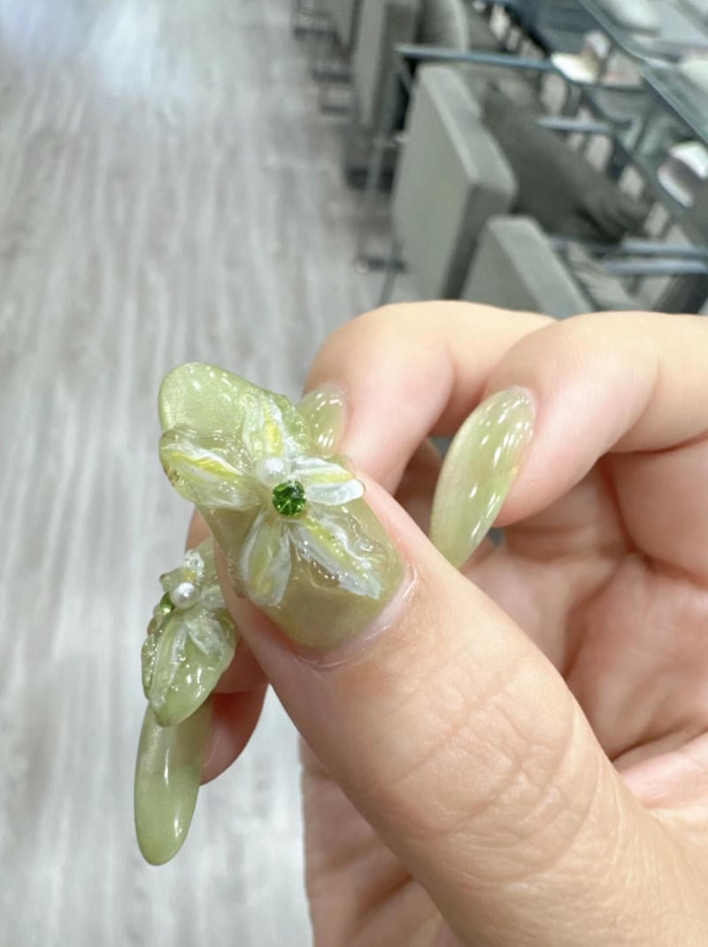 Móng tay được trang trí nghệ thuật với hoa nổi và đá quý màu xanh lá, nền phòng với sàn gỗ và ghế ngồi.